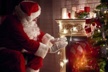 A Santa Claus figure in a red suit and white beard kneels beside a lit fireplace mantel with candles, warm lights, and a decorated Christmas tree in a cosy indoor holiday setting.