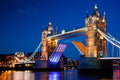 London Tower Bridge at night, illuminated in warm golden light with blue and white accents, with the bridge deck raised and reflections visible in the water under a deep blue sky.