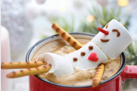 A marshmallow snowman decoration on a red mug filled with hot chocolate, garnished with pretzel sticks, set against a softly blurred festive background.