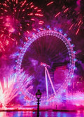 A vibrant night scene of the London Eye illuminated in pink and purple hues, surrounded by colorful fireworks exploding in the sky above the water.
